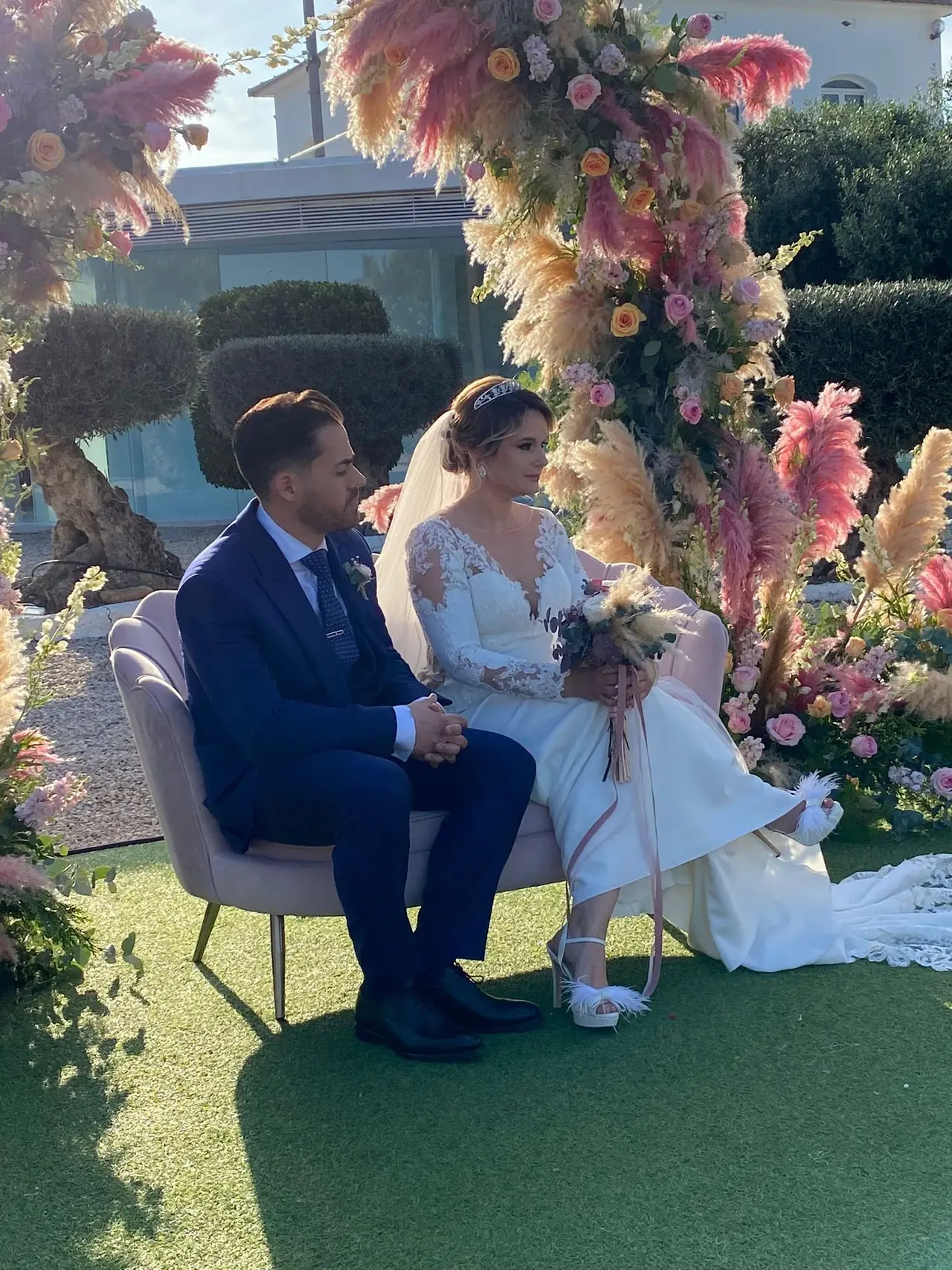 Pareja de novios sentados en banco de diseño elegante para exteriores durante la ceremonia de su boda.